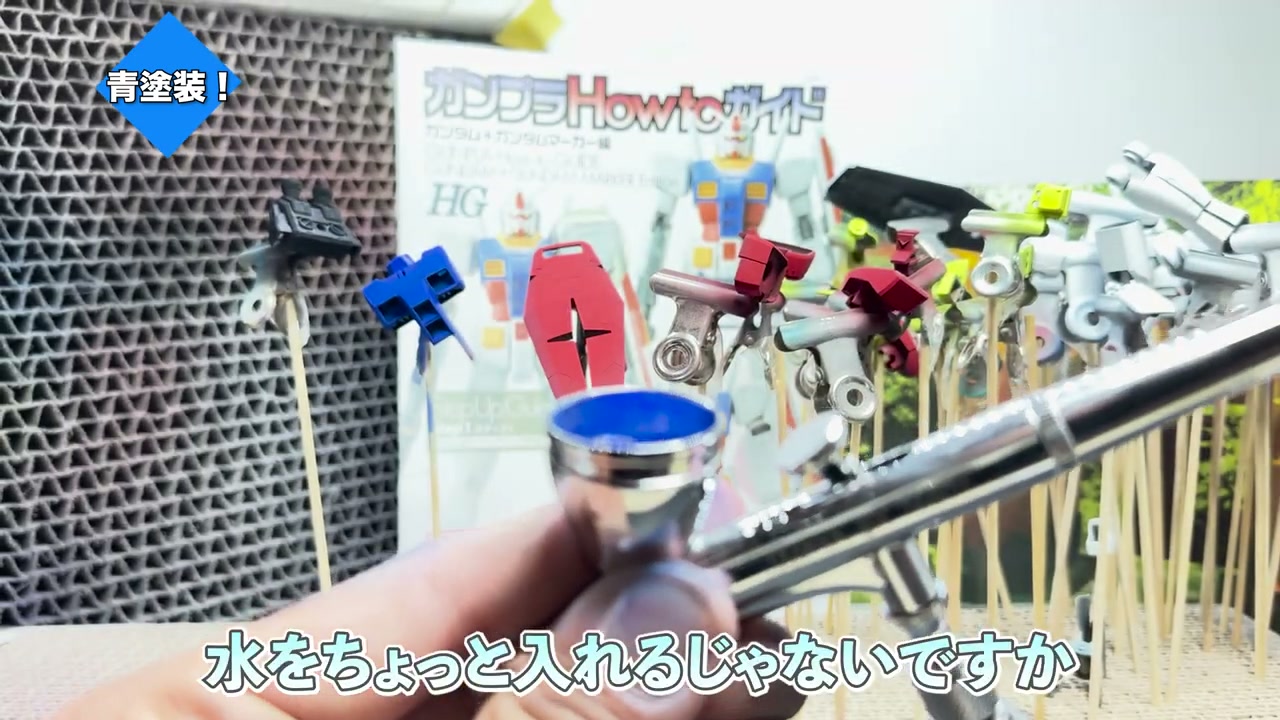 エアブラシで青い塗料を吹き付けている様子。エアブラシのカップに青塗料。「水をちょっと入れるじゃないですか」の字幕。